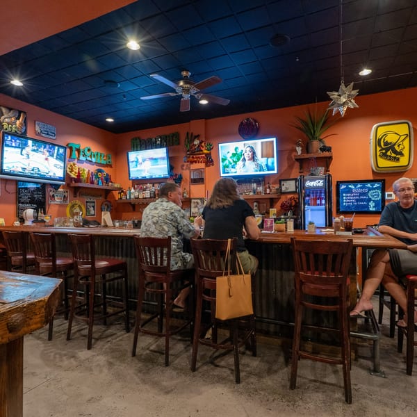Customers seated at a vibrant bar with multiple TVs, shelves of liquor, and Mexican decor.