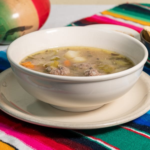 A white bowl of Mexican albondigas soup with meatballs, vegetables, and potatoes on a colorful striped placemat.