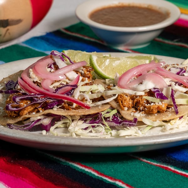 Two El Coronado tacos with shredded chicken, cabbage, pickled onions, and lime wedges.